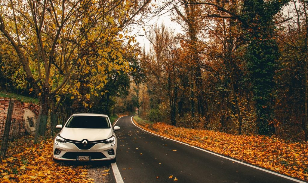 Une Renault Clio roulant sur une route d'automne, avec des feuilles brunes tombées au sol.